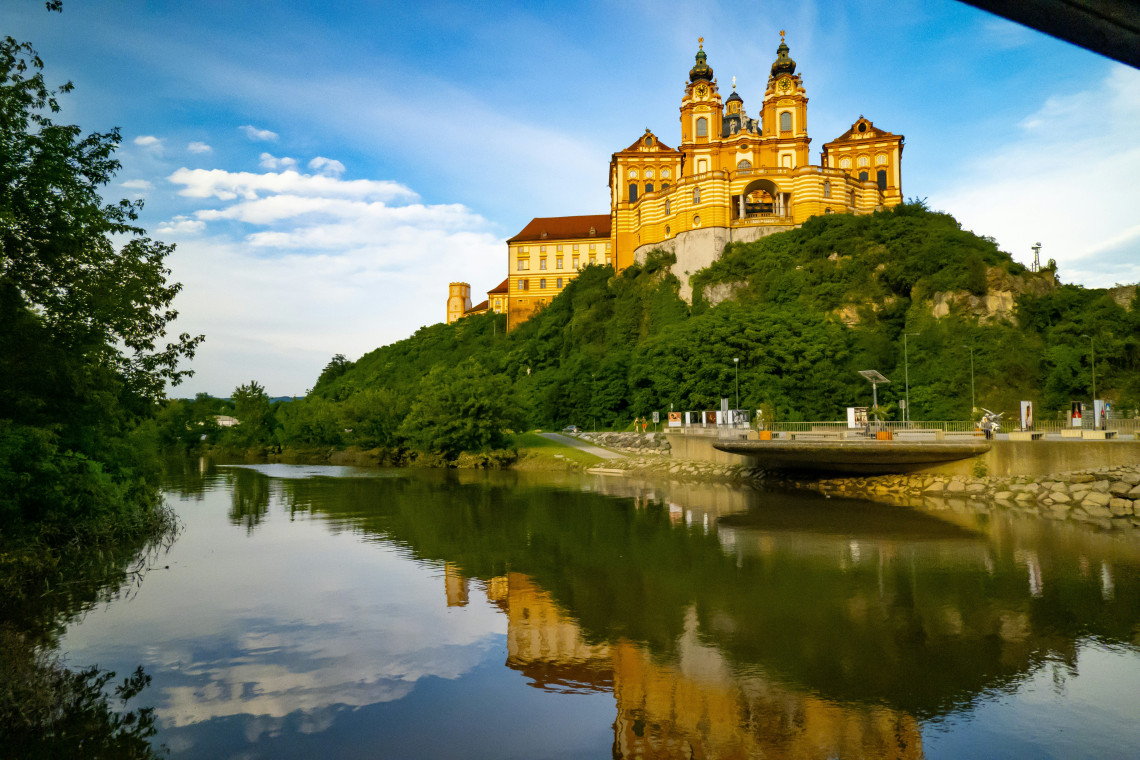 Melk Abbey rises above the Danube River in Melk, Austria, with its twin towers and yellow Baroque fa&ccedil;ade illuminated by late-afternoon light. The monastery and surrounding greenery are reflected in the water.
