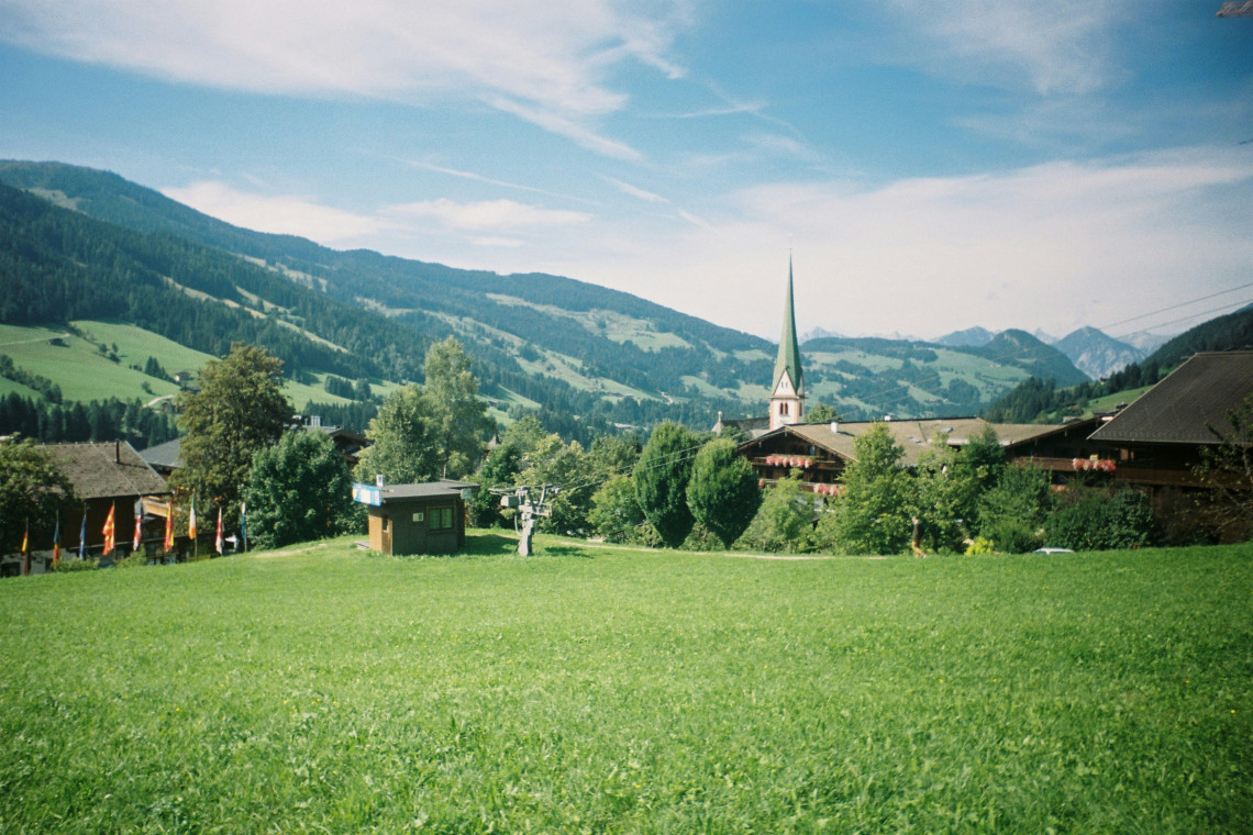 The village of Alpbach in Tyrol, Austria, is surrounded by green meadows, traditional wooden buildings and forested mountain slopes. A slender church spire rises above the rural Alpine landscape.