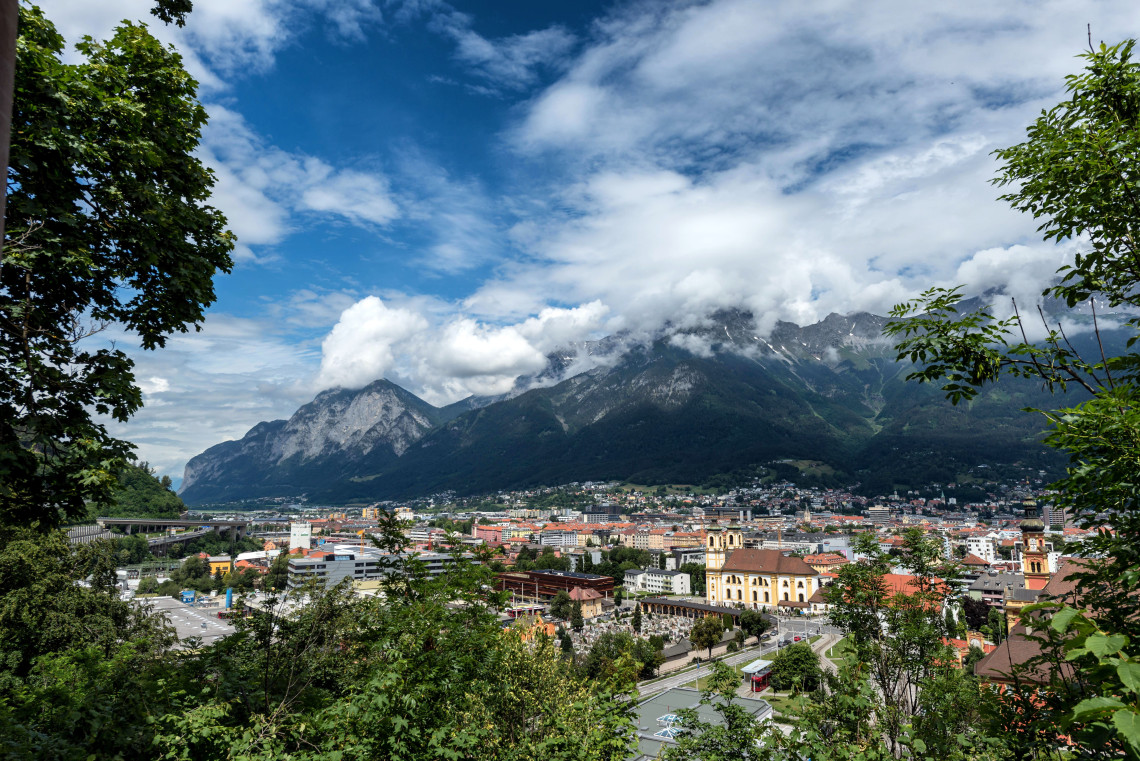 Panoramic view across Innsbruck, Austria, with the city spread beneath the steep Nordkette mountain range. Historic buildings, church towers and surrounding trees fill the valley under drifting clouds.