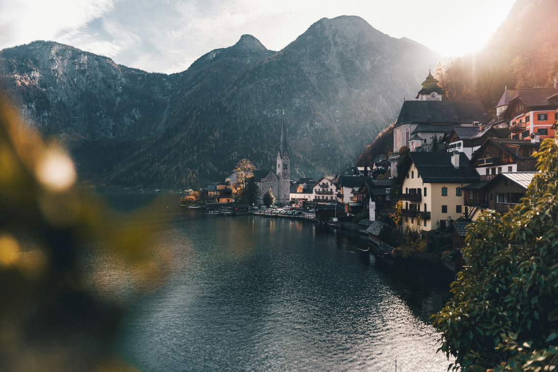 Hallstatt village stands beside Lake Hallstatt in Austria, with its historic church spire and traditional houses lining the waterfront. Steep forested mountains rise behind the village in warm sunlight.