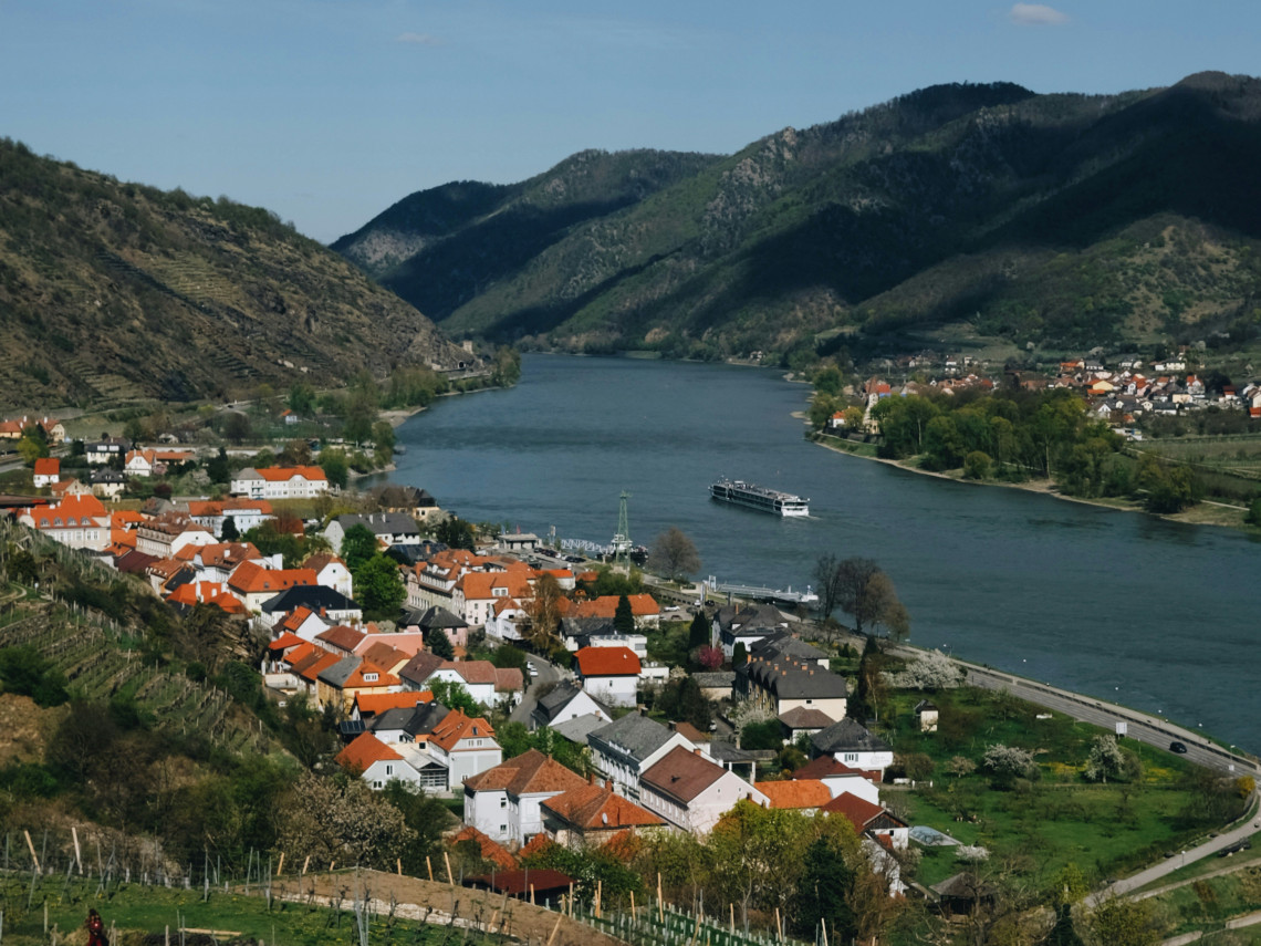 Spitz an der Donau lies beside the Danube River in Austria&rsquo;s Wachau Valley, surrounded by terraced vineyards and wooded hills. A river cruise ship passes the village&rsquo;s red-roofed houses along the broad waterway.