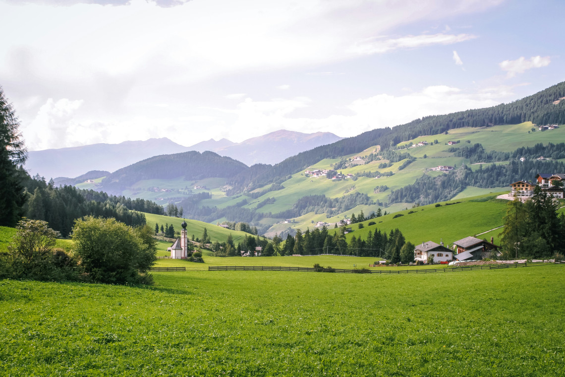 Rolling green meadows, scattered farmhouses and a small church fill a rural valley in Tyrol, Austria. Forested hills and layered Alpine mountains extend into the distance beneath a bright sky.
