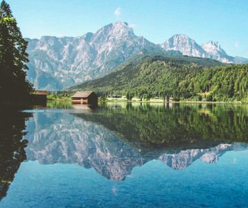 Almsee in Grünau im Almtal, Austria, reflects the forested shoreline and peaks of the Totes Gebirge mountain range. A small lakeside boathouse stands beside the calm Alpine water.