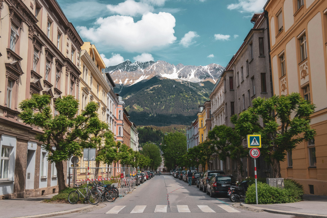 A residential street in Innsbruck, Austria, is lined with historic buildings, trees, parked cars and bicycles. The snow-dusted Nordkette mountains rise directly beyond the city.