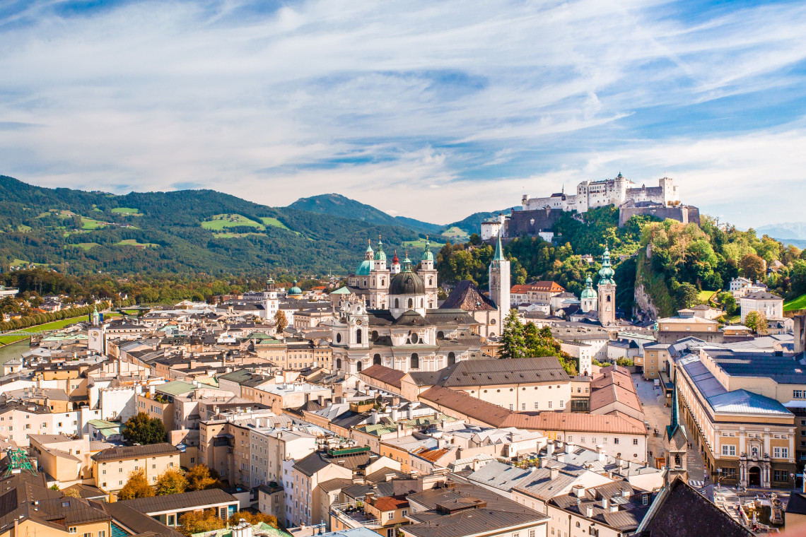 Panoramic view across Salzburg Old Town in Salzburg, Austria, with church domes and historic rooftops below Hohensalzburg Fortress. Green hills surround the city beneath a partly cloudy sky.