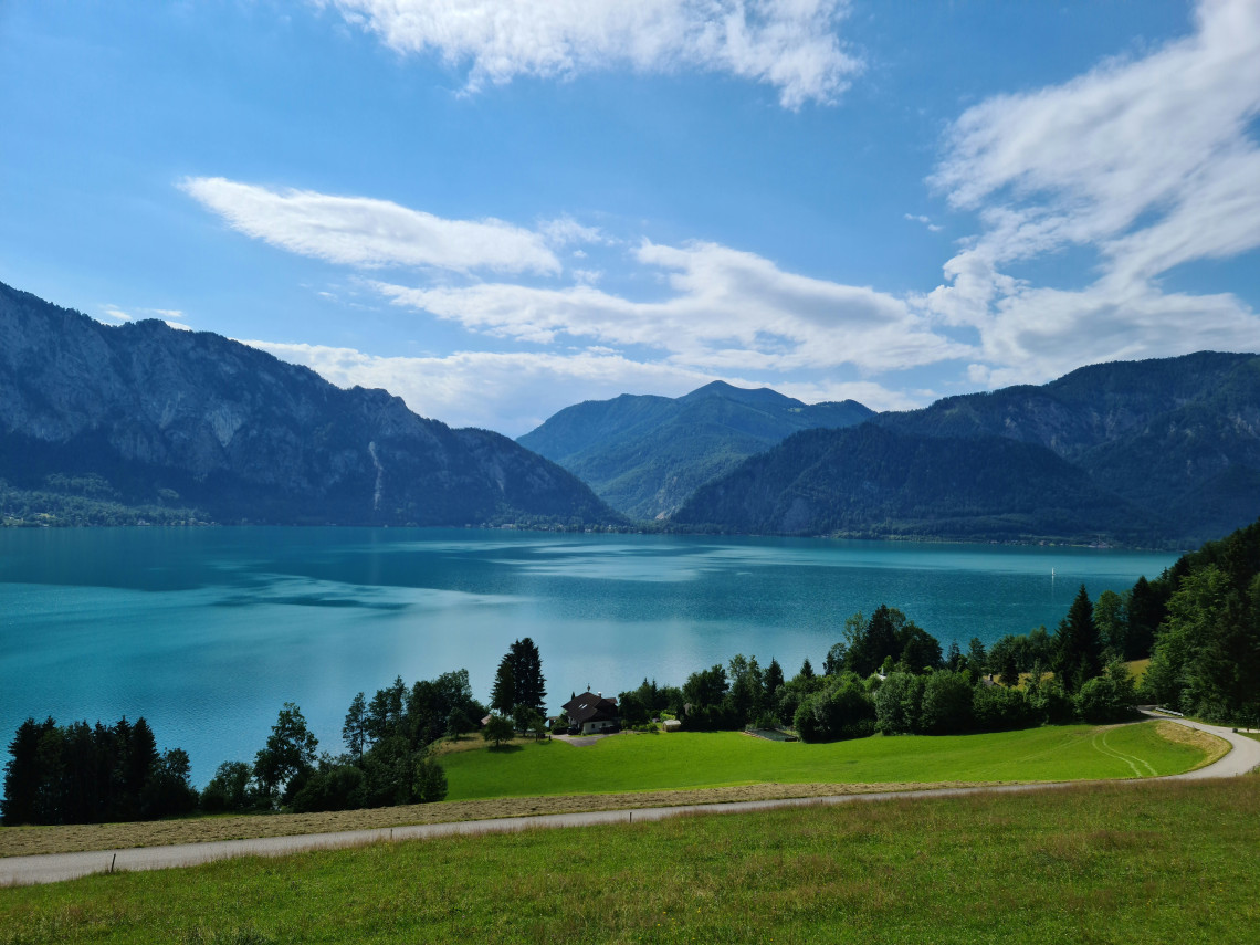 Lake Wolfgang stretches between forested mountains in Austria&rsquo;s Salzkammergut region. Green meadows, scattered houses and a winding country road overlook the blue Alpine water.