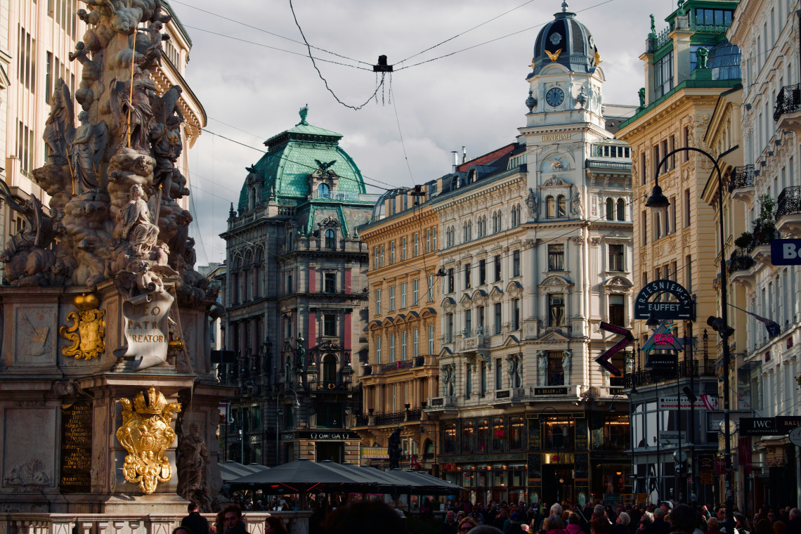 The Baroque Plague Column stands along the Graben in central Vienna, Austria, surrounded by ornate historic buildings, shops and pedestrians. Tram wires cross above the busy city-centre street.