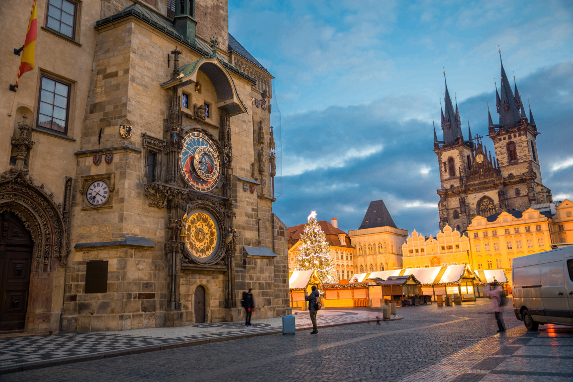 The Prague Astronomical Clock overlooks Christmas market stalls and a decorated tree in Old Town Square, Prague, Czech Republic. The Church of Our Lady before T&yacute;n rises behind the historic square at dusk.