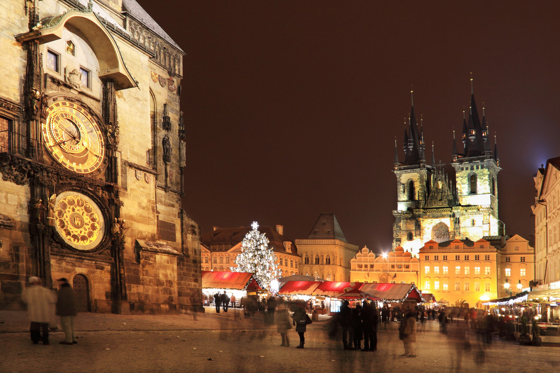 Night view of Old Town Square Christmas Market in Prague, Czech Republic, with the illuminated Astronomical Clock, seasonal stalls and a Christmas tree. The twin towers of the Church of Our Lady before T&yacute;n frame the market.
