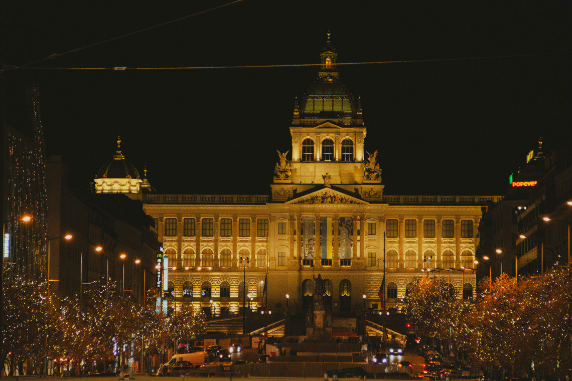 The National Museum is illuminated above Wenceslas Square in Prague, Czech Republic, with rows of trees decorated in seasonal lights. Evening traffic and pedestrians move through the city-centre boulevard.
