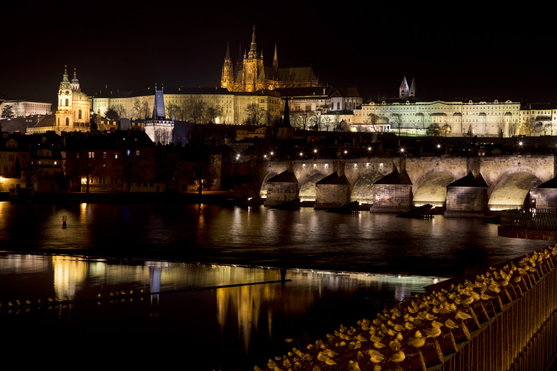 Charles Bridge crosses the Vltava River beneath the illuminated Prague Castle complex in Prague, Czech Republic. Historic buildings and stone arches reflect in the dark water at night.