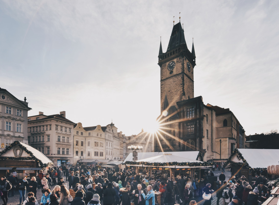 Visitors gather among Christmas market stalls beneath the Old Town Hall tower in Prague, Czech Republic. Low winter sunlight passes between the historic buildings of Old Town Square.