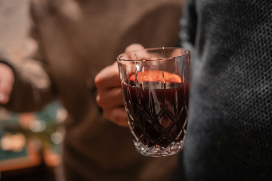 A visitor holds a clear glass of dark red mulled wine at a Christmas market in Prague, Czech Republic. Other market guests appear softly blurred in the background.