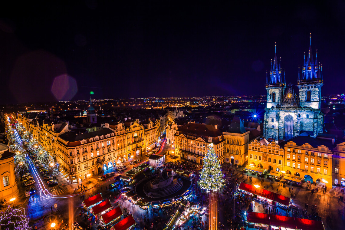 Aerial night view of Old Town Square Christmas Market in Prague, Czech Republic, with red-roofed stalls, a large illuminated Christmas tree and surrounding historic buildings. The Church of Our Lady before T&yacute;n dominates the festive city-centre scene.
