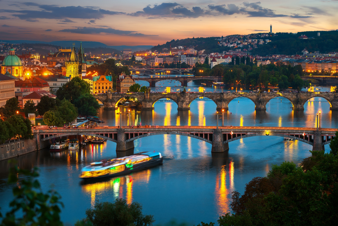 Elevated sunset view across the Vltava River in Prague, Czech Republic, showing Charles Bridge and several illuminated bridges extending through the historic city. Boats and riverside landmarks reflect in the water beneath the evening skyline.