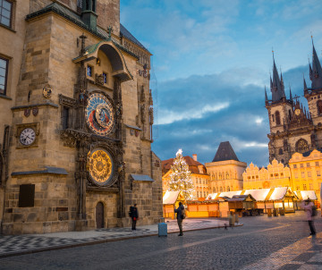 The Prague Astronomical Clock stands beside illuminated Christmas market stalls in Old Town Square, Prague, Czech Republic. The Church of Our Lady before Týn and historic buildings rise in the background at dusk.