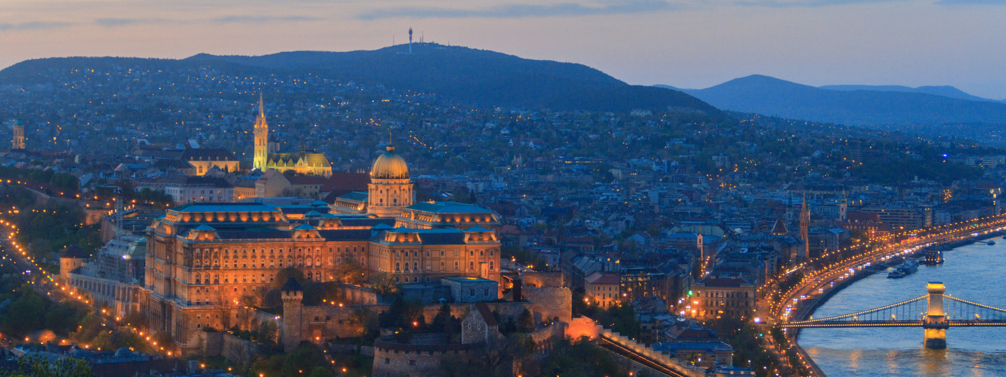 Panoramic evening view of Buda Castle overlooking the Danube River in Budapest, Hungary. The illuminated palace complex, riverside roads and Széchenyi Chain Bridge extend across the cityscape.