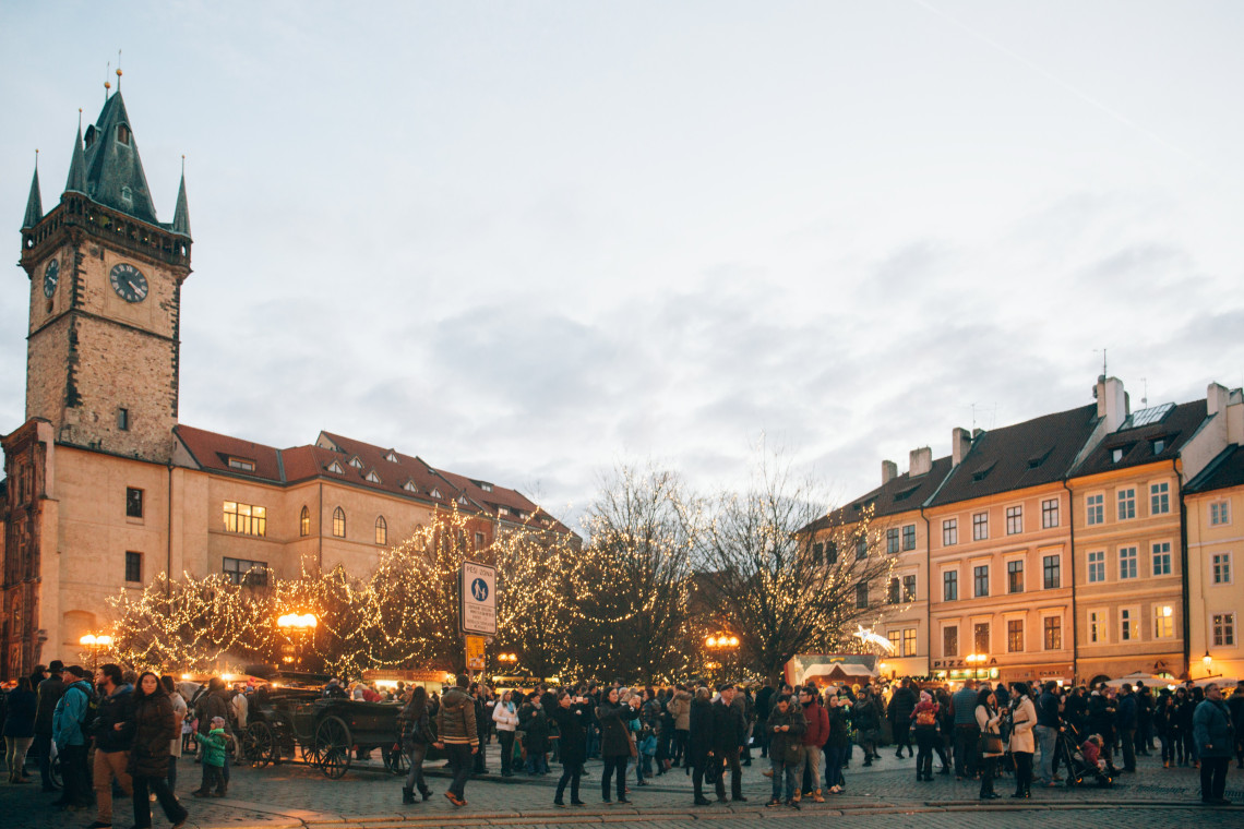 Visitors gather among illuminated trees and seasonal stalls in Old Town Square, Prague, Czech Republic. The medieval Old Town Hall tower rises above the Christmas market and surrounding historic buildings.