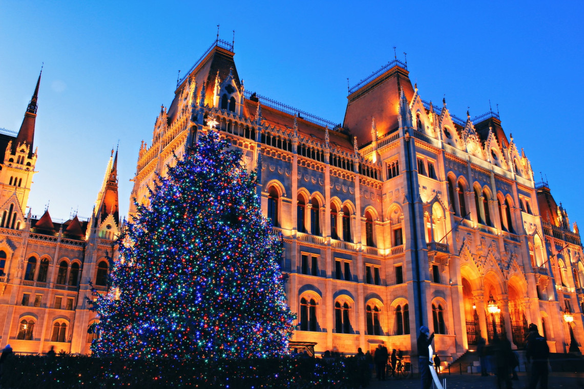 A large Christmas tree covered in colourful lights stands beside the Hungarian Parliament Building in Budapest, Hungary. The illuminated Neo-Gothic fa&ccedil;ade rises behind the seasonal display at dusk.