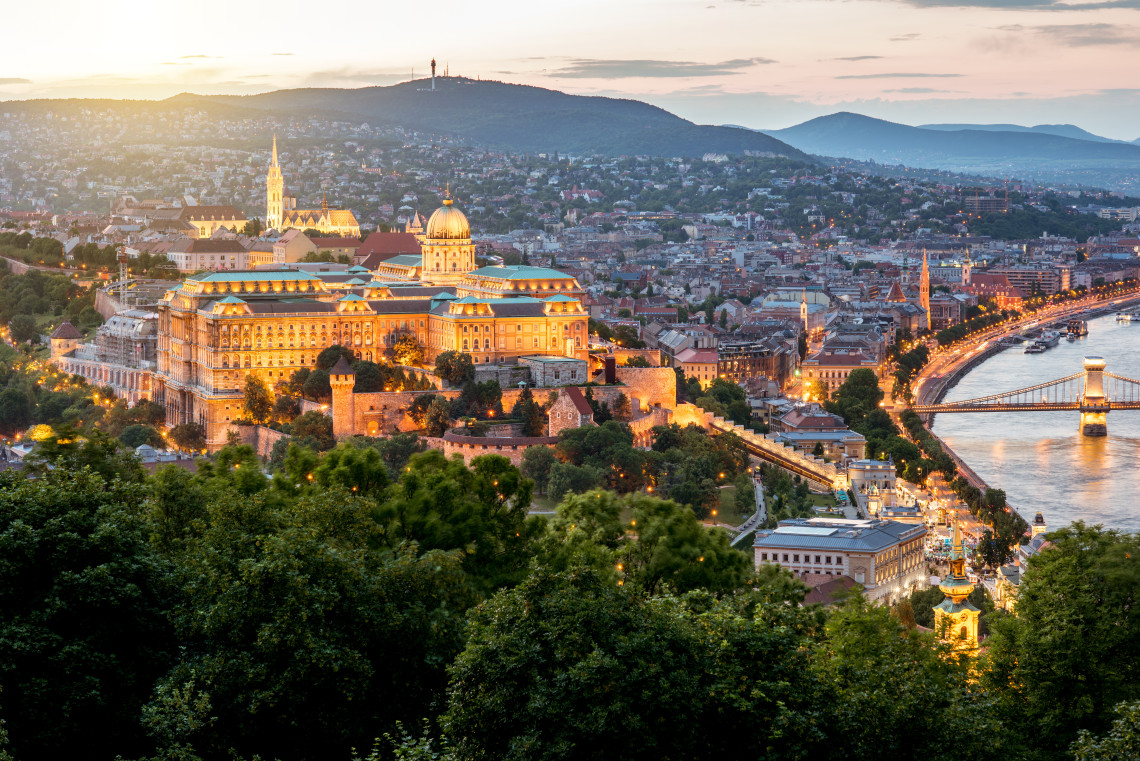 Panoramic sunset view of Buda Castle overlooking the Danube River in Budapest, Hungary. The illuminated palace complex, riverside streets and Sz&eacute;chenyi Chain Bridge extend across the cityscape.