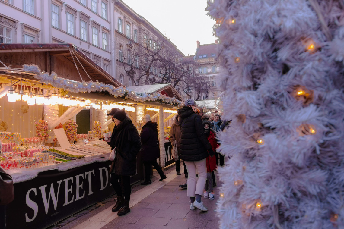 A Christmas market stall in Budapest, Hungary, displays colourful sweets and candy beneath warm lights. Visitors walk between decorated wooden stalls beside a white illuminated Christmas tree.