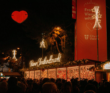 Visitors gather beneath the illuminated entrance to the Wiener Christkindlmarkt at Rathausplatz in Vienna, Austria. A red heart decoration, seasonal lights and festive market stalls fill the evening scene.