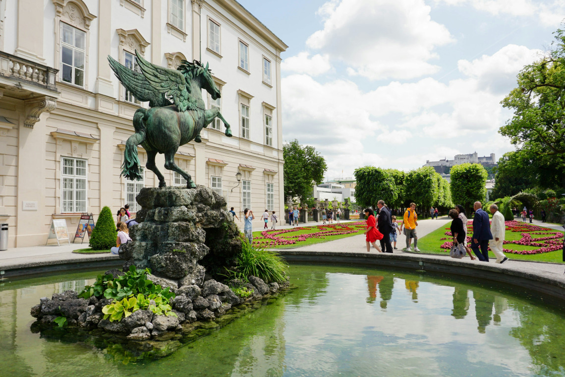 The bronze Pegasus statue stands above a fountain in Mirabell Gardens, Salzburg, Austria. Formal flowerbeds, trimmed hedges and visitors surround the Baroque garden landmark, with Hohensalzburg Fortress visible in the distance.