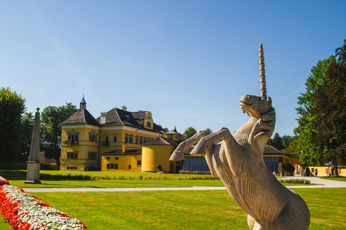 A decorative stone figure stands within the landscaped grounds of Hellbrunn Palace in Salzburg, Austria. The yellow palace buildings and wooded gardens appear beneath a clear blue sky.