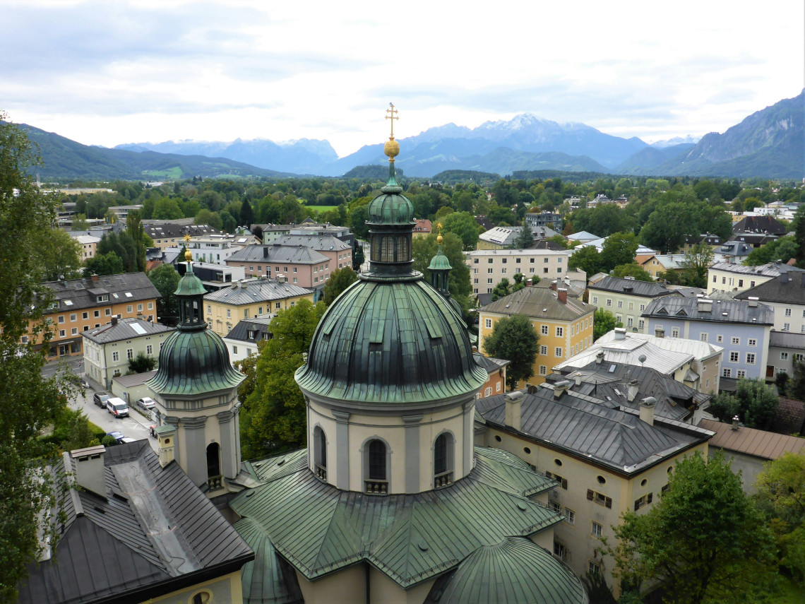 Elevated view of Salzburg Cathedral in Salzburg, Austria, showing its large copper dome and surrounding historic rooftops. Alpine mountains rise beyond the city beneath a cloudy sky.