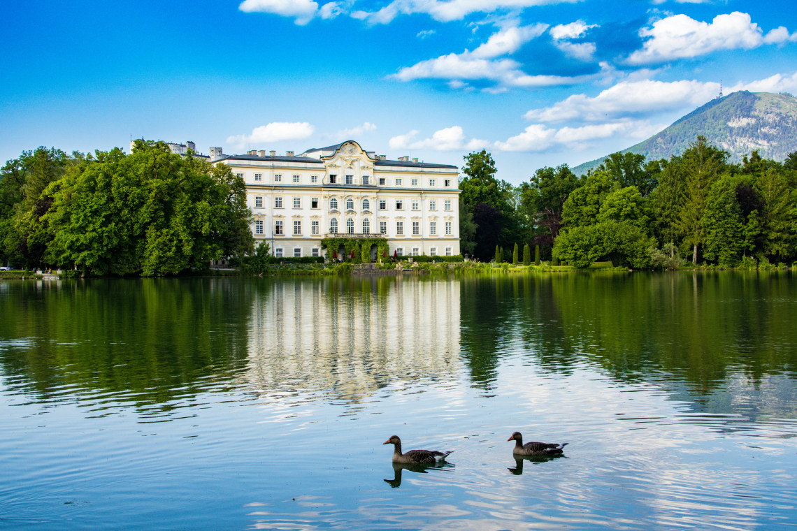 Leopoldskron Palace stands beside its reflective lake in Salzburg, Austria, surrounded by mature trees and landscaped grounds. Ducks cross the water with the Alpine foothills visible behind the Baroque residence.