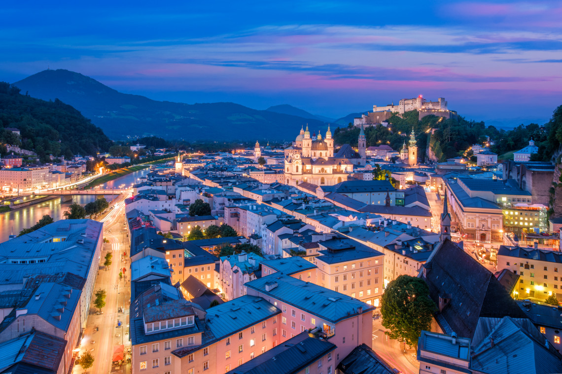 Panoramic dusk view of Salzburg Old Town in Austria, with illuminated churches, historic rooftops and the Salzach River extending through the city. Hohensalzburg Fortress rises above the skyline under a blue and pink evening sky.