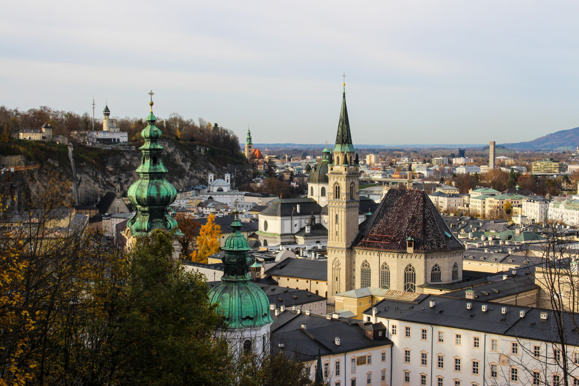Historic church towers rise above the rooftops of Salzburg Old Town in Austria. The surrounding cityscape extends toward wooded hills and modern districts under a pale sky.