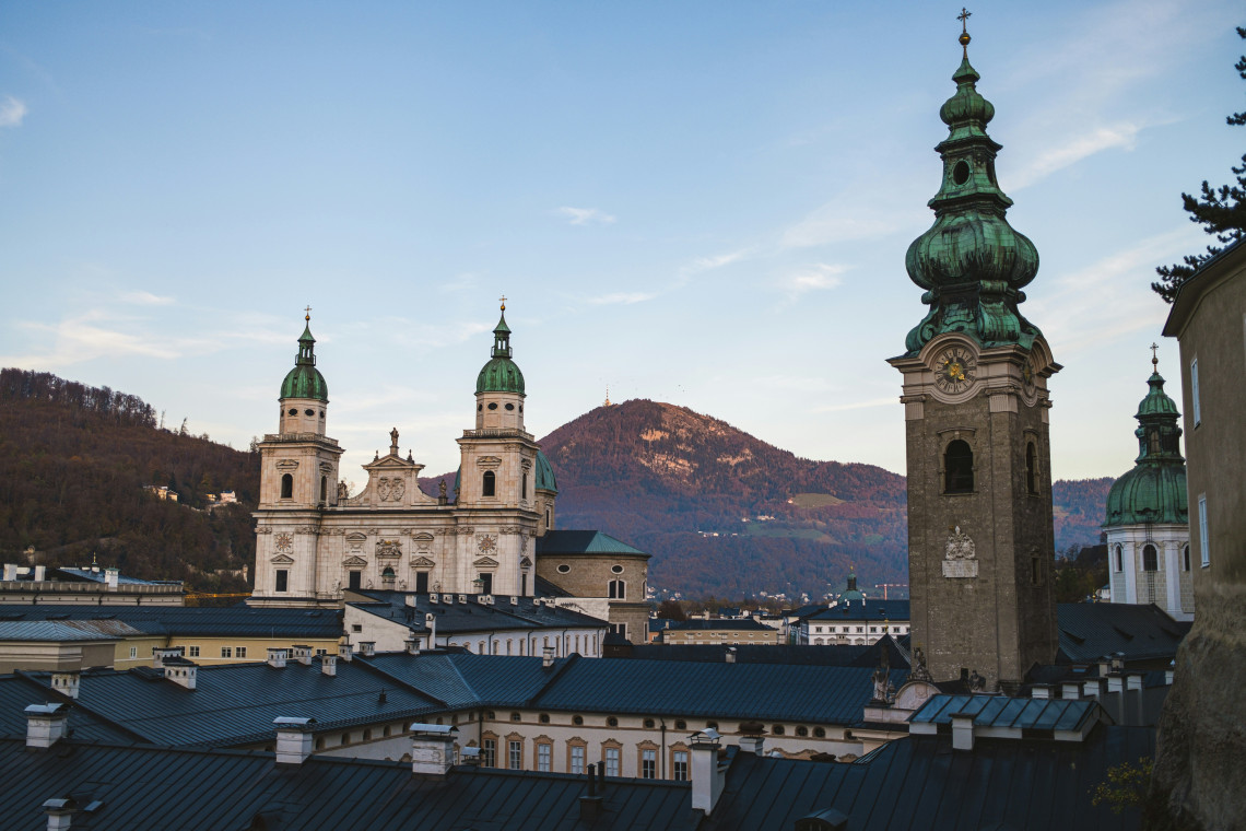 Salzburg Cathedral and neighbouring Baroque church towers rise above the historic centre of Salzburg, Austria. Dark rooftops and wooded hills surround the landmarks in the late afternoon light.