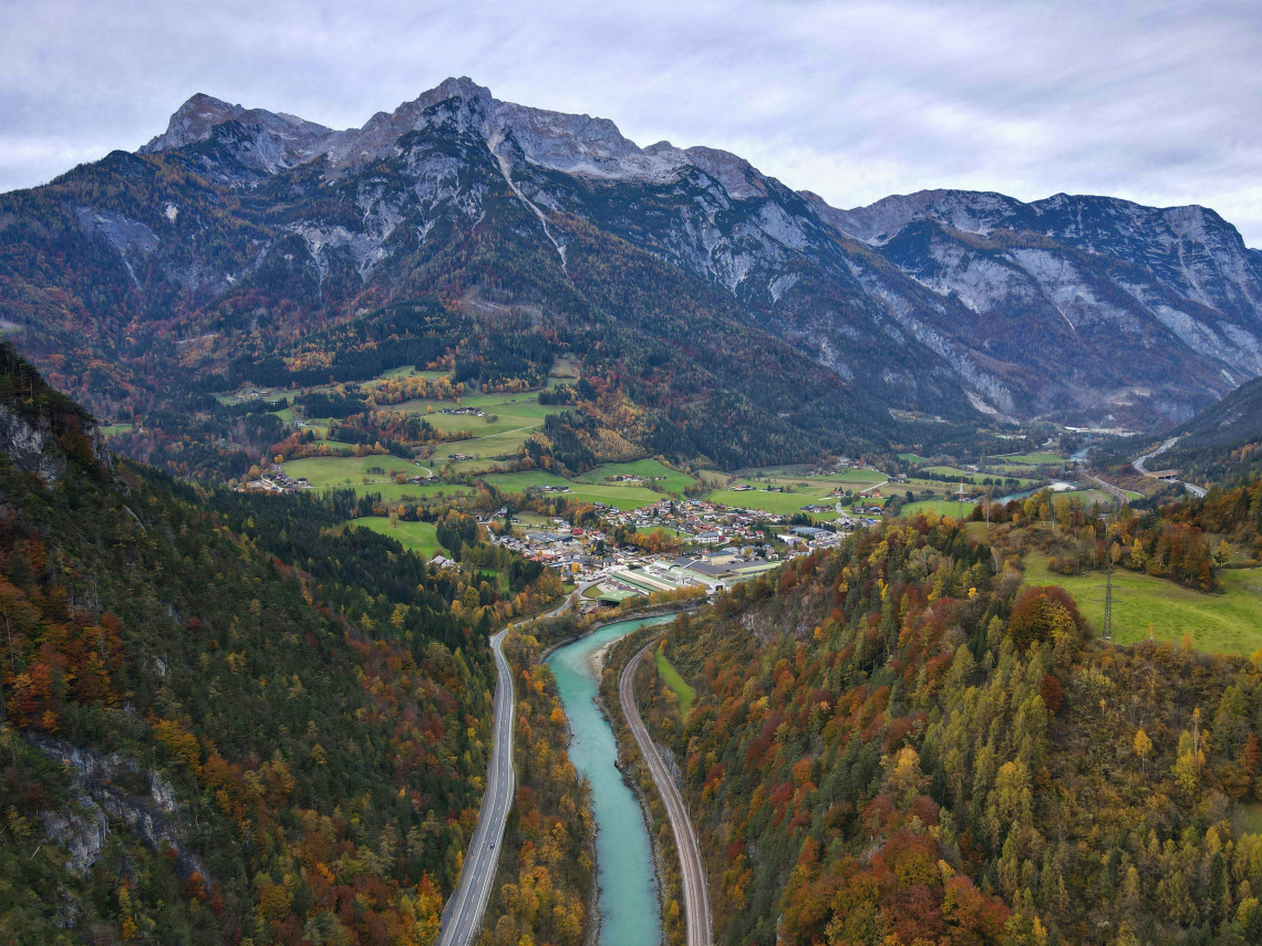 The Salzach River runs through a broad Alpine valley in Salzburg, Austria, beside roads, railway tracks and a small settlement. Forested slopes and rocky mountains display autumn colours across the landscape.