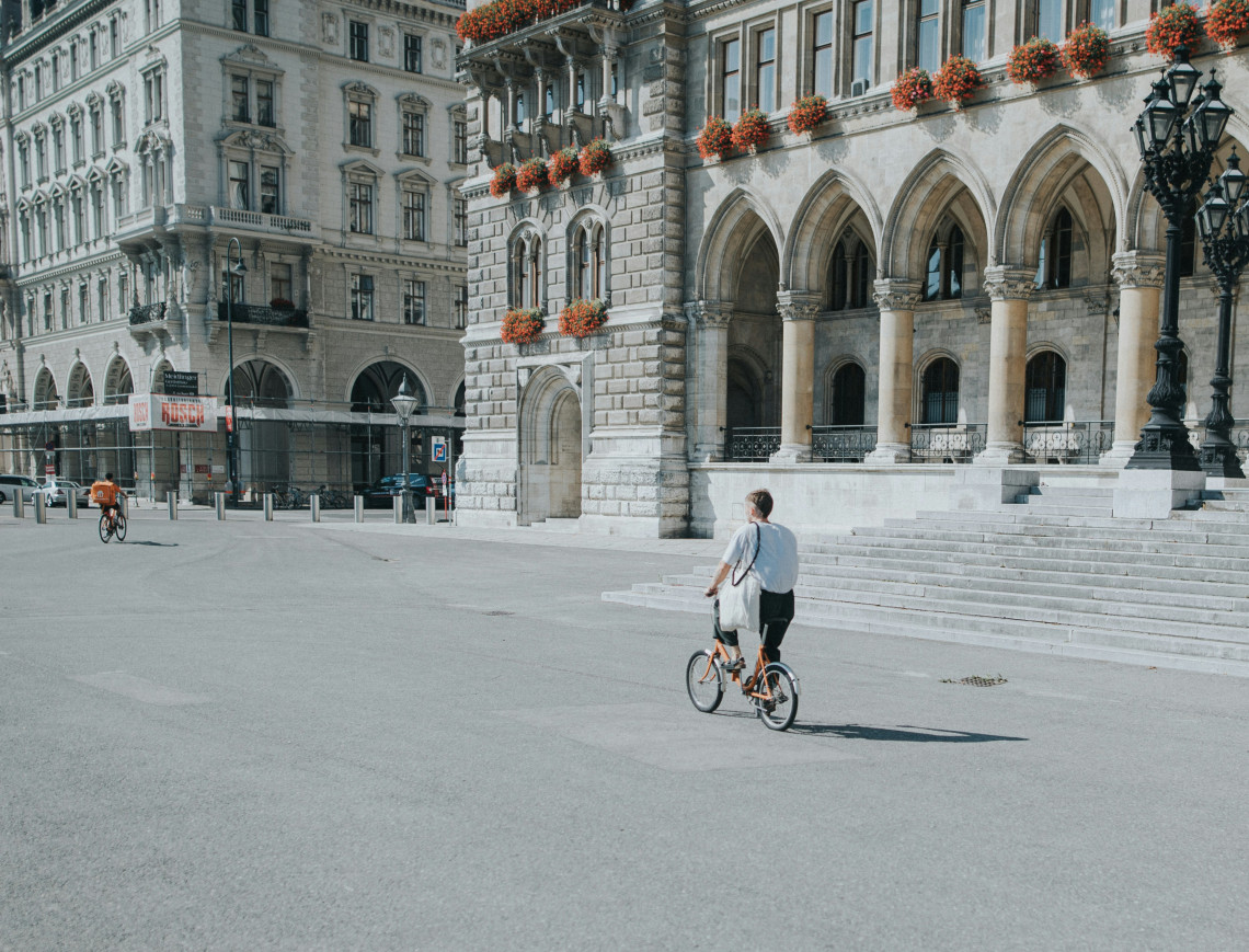 A cyclist crosses Rathausplatz beside the Neo-Gothic arcades of Vienna City Hall in Vienna, Austria. Stone fa&ccedil;ades, flower-filled windows and historic street lamps frame the open square.