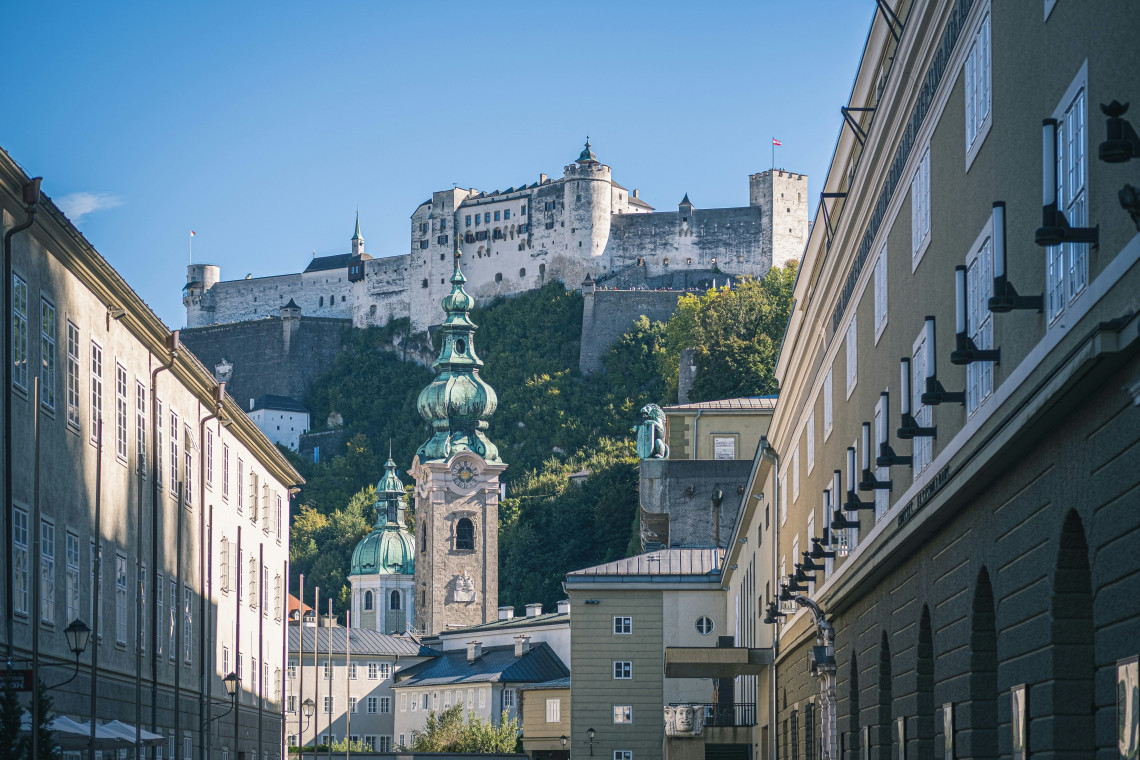 Hohensalzburg Fortress rises above a narrow street in Salzburg, Austria, with a green-domed Baroque church tower in the foreground. Historic buildings frame the hilltop fortress beneath a clear blue sky.