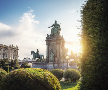 The Maria Theresa Monument rises among manicured hedges at Maria-Theresien-Platz in Vienna, Austria. Evening sunlight illuminates the monumental sculpture and surrounding imperial architecture.