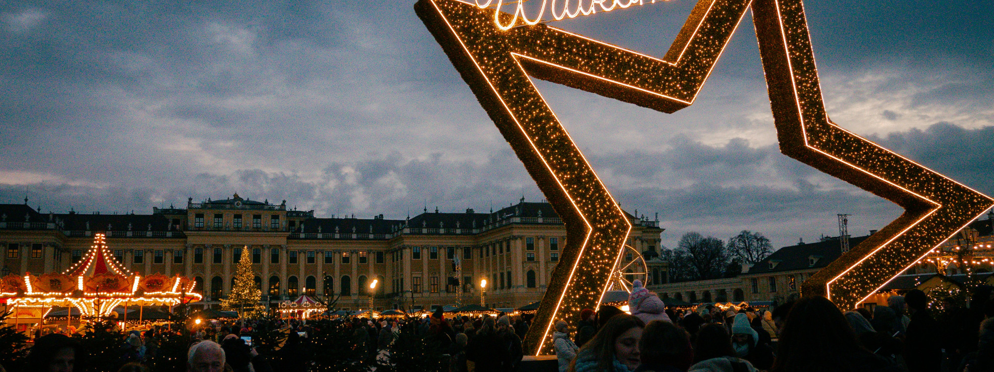 A large illuminated star frames the Christmas market in front of Schönbrunn Palace in Vienna, Austria. Visitors gather among festive stalls and seasonal lights beneath an overcast evening sky.