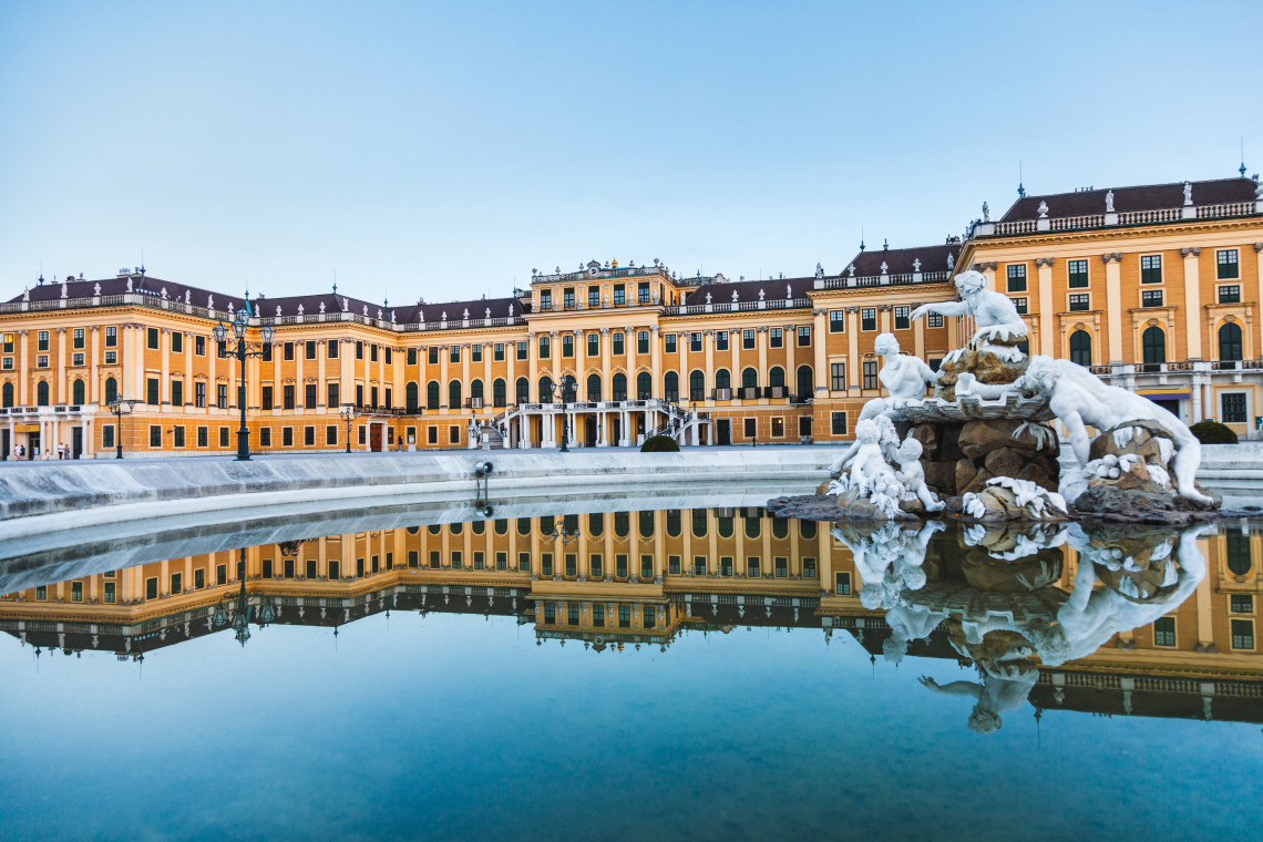 Winter view of Sch&ouml;nbrunn Palace in Vienna, Austria, with snow covering the Neptune Fountain and surrounding grounds. The yellow Baroque palace is reflected in the still water.