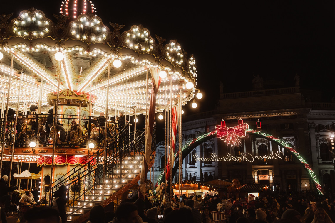 A brightly illuminated two-level carousel welcomes visitors to the Wiener Christkindlmarkt at Rathausplatz in Vienna, Austria. Crowds gather beneath festive lights and the market&rsquo;s decorated entrance arch.