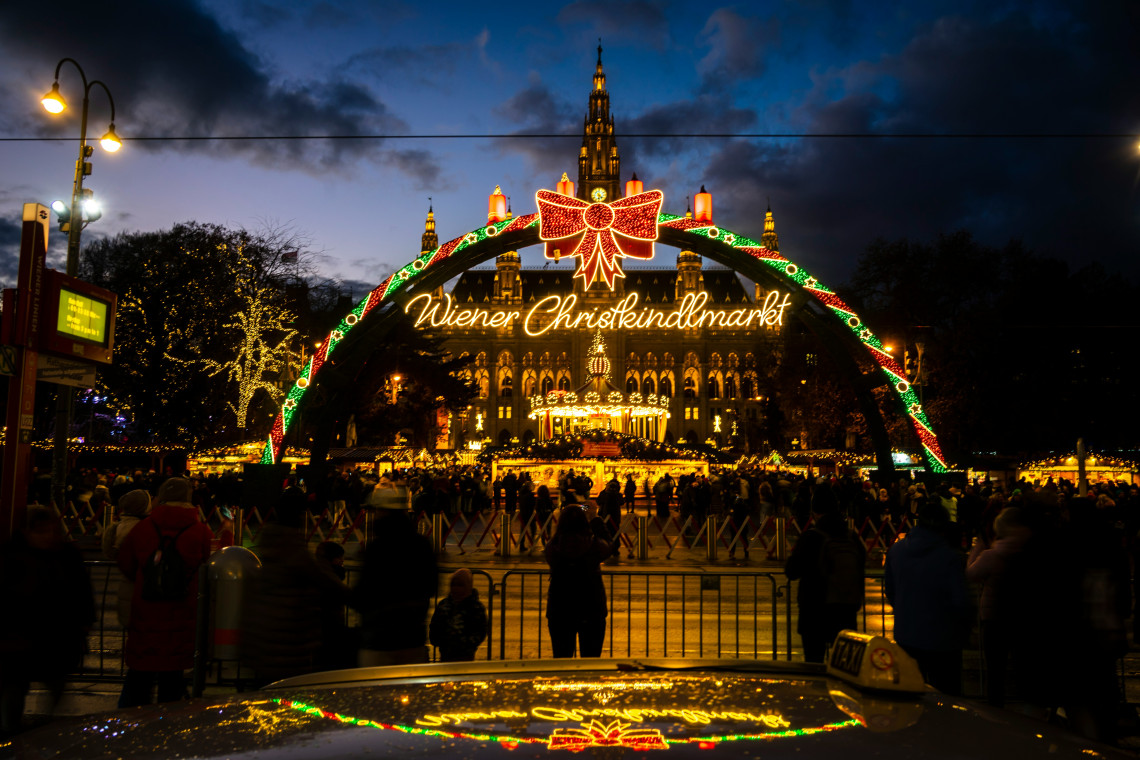 The illuminated entrance to the Wiener Christkindlmarkt frames Vienna City Hall at Rathausplatz in Vienna, Austria. Visitors gather around the seasonal market beneath Christmas lights and a large red bow.