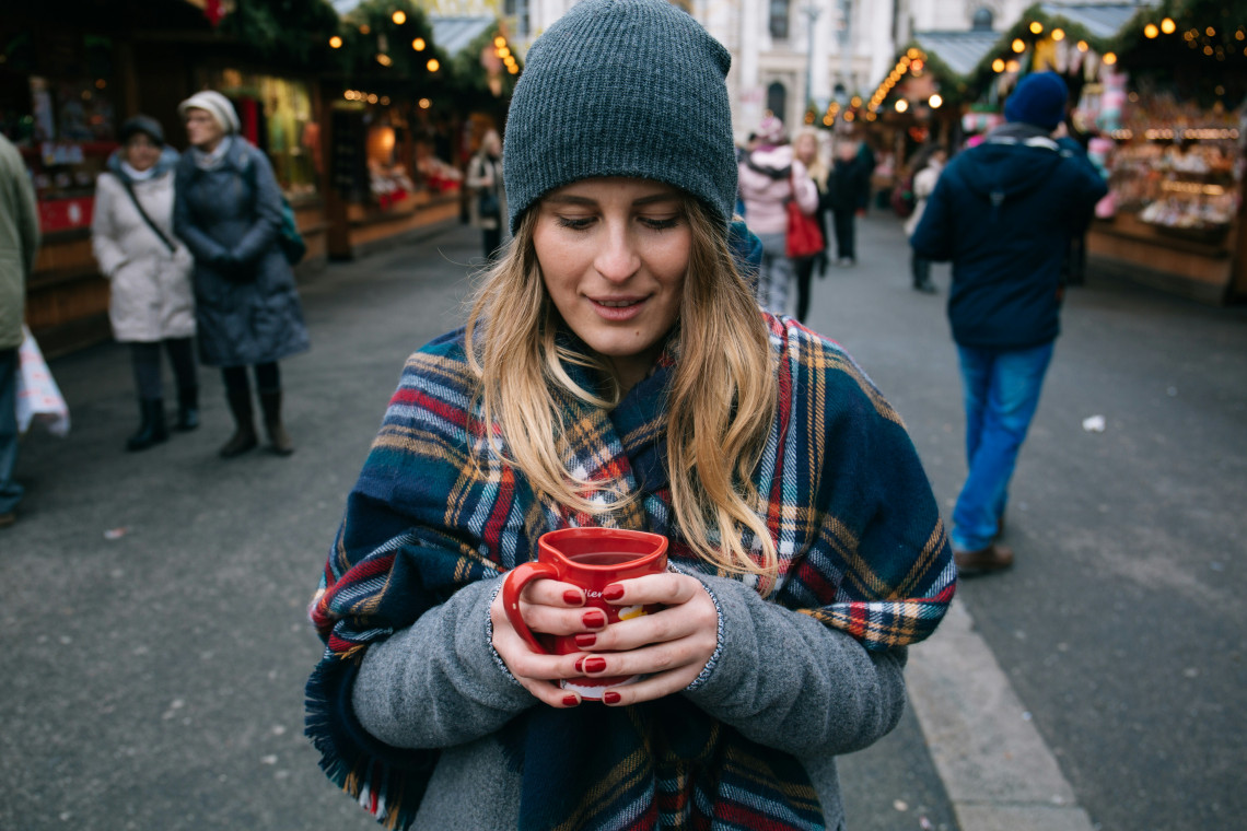 A visitor holds a red ceramic cup of warm Gl&uuml;hwein at a Christmas market in Vienna, Austria. Wooden stalls, seasonal decorations and other market visitors appear in the background.