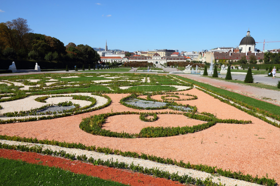 Formal gardens at the Belvedere complex in Vienna, Austria, feature geometric planting beds, gravel pathways and carefully shaped greenery. The Vienna skyline and the domed Salesian Church appear beyond the landscaped grounds.