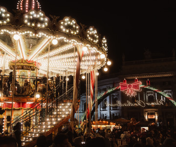 An illuminated vintage carousel surrounded by visitors and Christmas market stalls at Schönbrunn Palace in Vienna, Austria. Festive lights and decorations fill the market after dark.