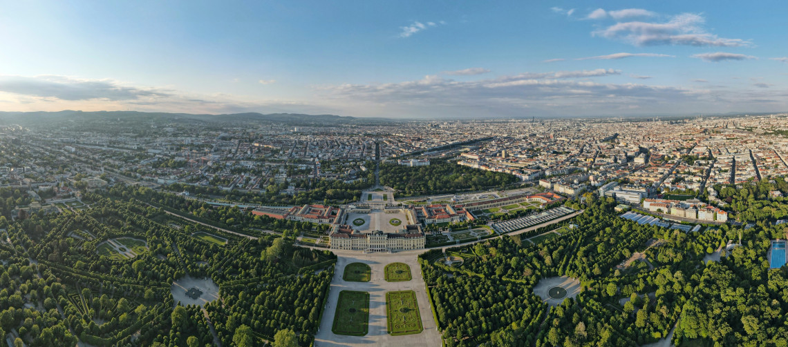 Aerial view of Sch&ouml;nbrunn Palace and its formal gardens in Vienna, Austria. Geometric lawns, tree-lined avenues and the yellow Baroque palace extend toward the city skyline.