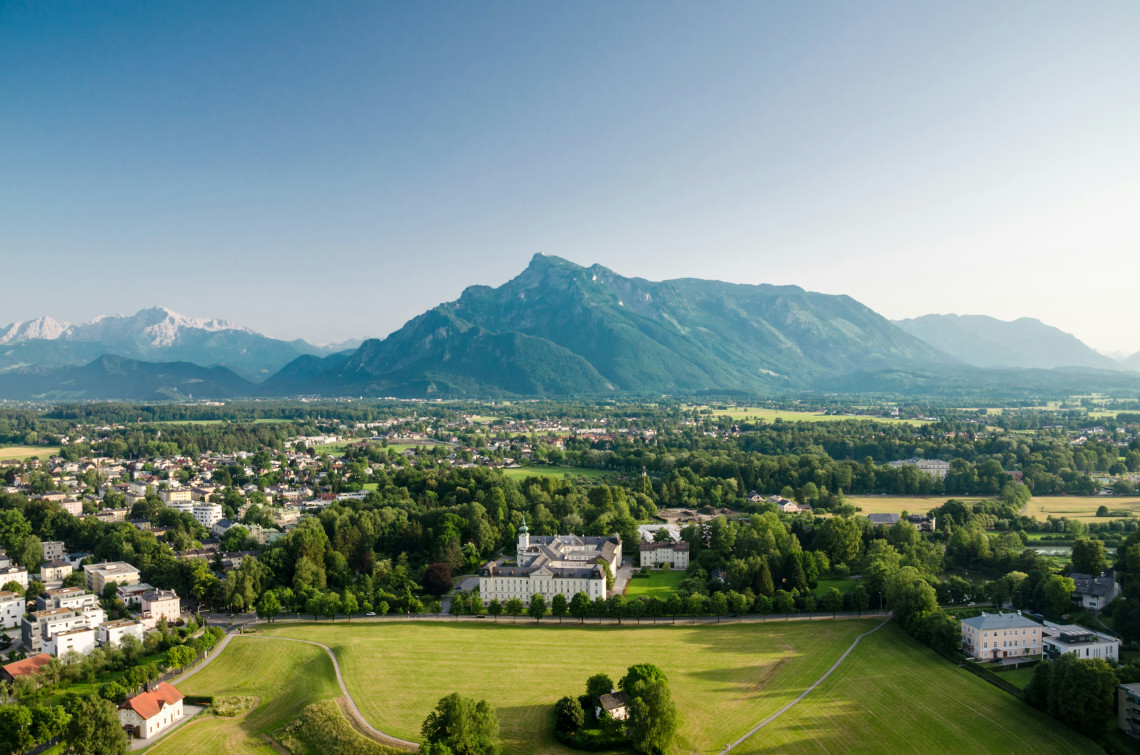 Panoramic view across Salzburg, Austria, with residential neighbourhoods, green parkland and historic buildings extending toward Untersberg Mountain. Forested foothills and Alpine peaks define the distant skyline.
