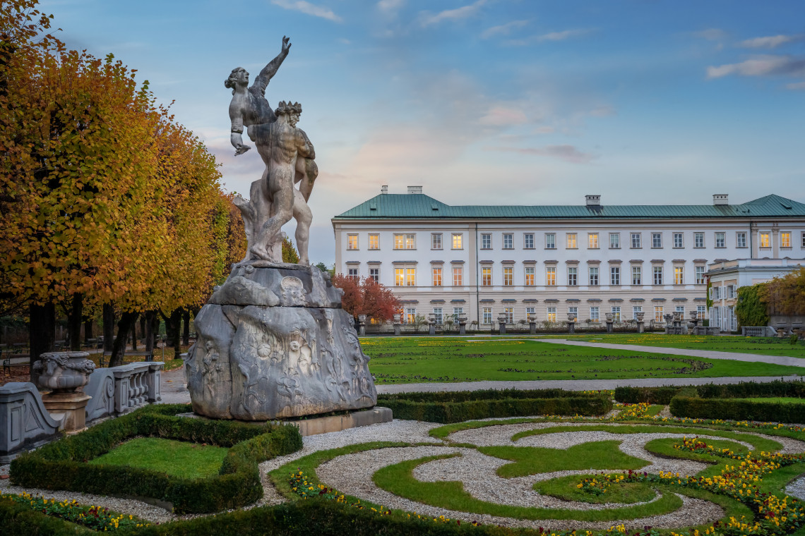 A large Baroque sculpture stands among geometric flowerbeds and trimmed hedges in Mirabell Gardens, Salzburg, Austria. Mirabell Palace forms the backdrop beneath an autumn sky.