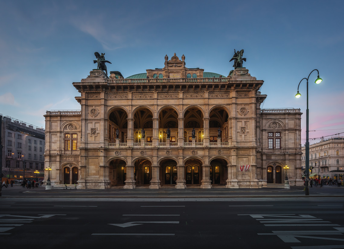 The Vienna State Opera stands illuminated beside the Ringstrasse in Vienna, Austria. Arched loggias, statues and its detailed Neo-Renaissance fa&ccedil;ade are visible beneath the evening sky.