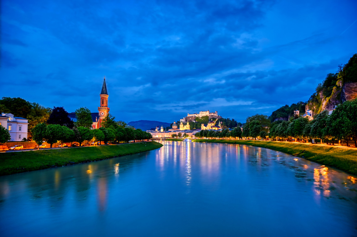 The Salzach River flows through Salzburg, Austria, beneath an illuminated skyline featuring Hohensalzburg Fortress and historic church towers. Riverside lights reflect across the water during blue hour.