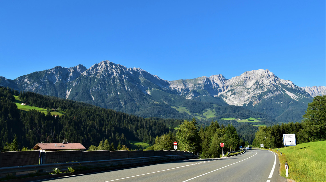 A paved road curves through green countryside beneath the Wilder Kaiser mountain range in Tyrol, Austria. Forested slopes and exposed limestone peaks rise under a clear blue sky.